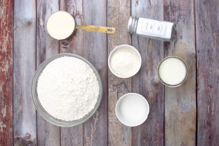 Various baking ingredients for english muffins recipe displayed on a wooden surface, including flour, sugar, baking powder, and milk.