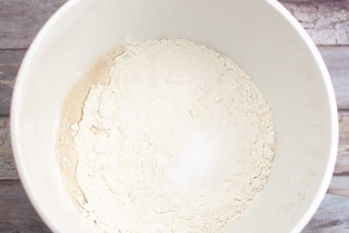 White mixing bowl containing dry flour on a wooden surface.