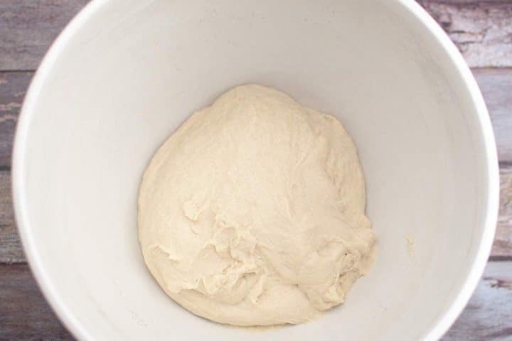 A ball of raw dough resting inside a white mixing bowl placed on a wooden surface.