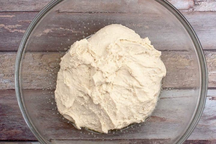 Raw bread dough in a clear glass bowl on a wooden table.