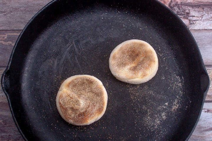Two english muffins toasting in a black cast iron skillet on a wooden surface.
