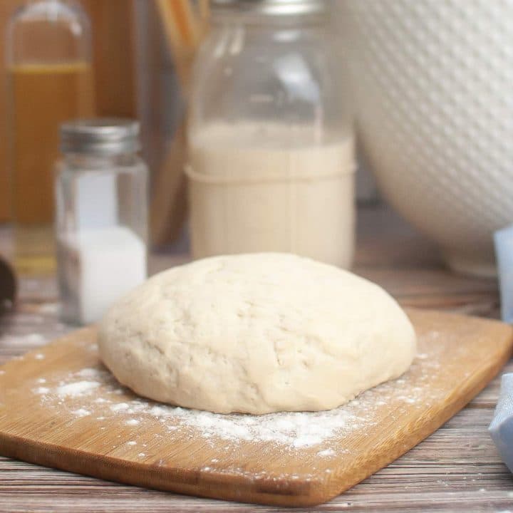 Fresh sourdough pizza dough on a wooden board with flour, accompanied by baking ingredients in the background.