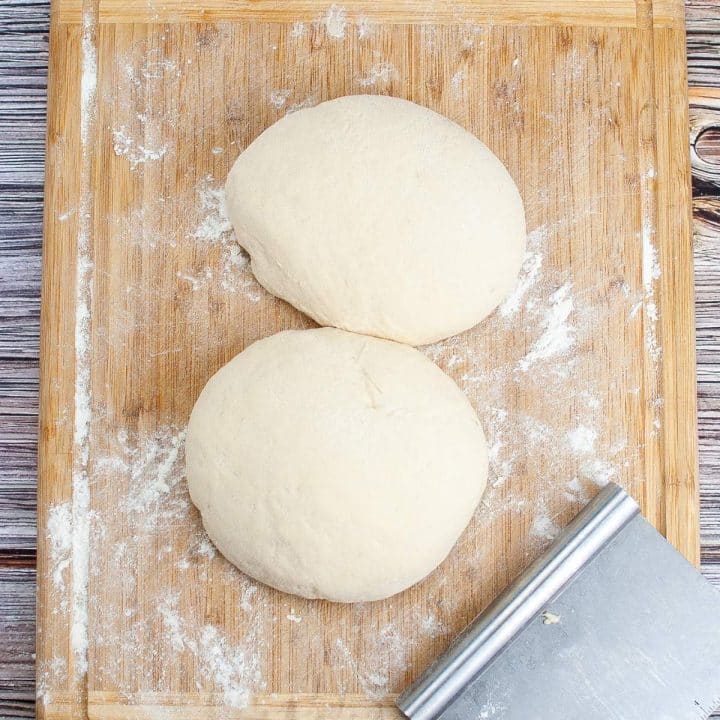 Two balls of dough on a wooden board with a dusting of flour and a dough scraper alongside them.