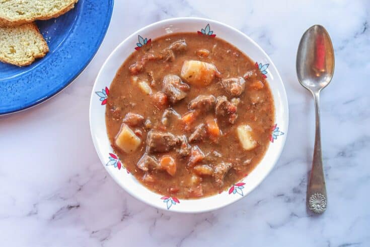 A bowl of beef stew with chunks of meat, potatoes, and carrots sits next to a spoon on a white marble surface, with a plate of bread partially visible—perfect inspiration for venison dinner ideas.